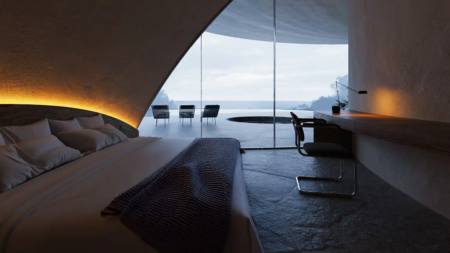 A view from a low, cave-like bed covered in textured blankets, past a writing desk with a lamp, towards floor-to-ceiling glass windows that open to a foggy terrace with chairs and the ocean beyond, framed by an asymmetrical concrete arch.