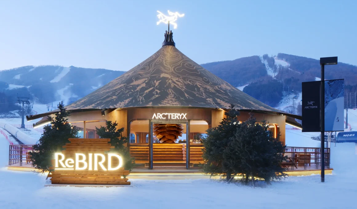 Eye-level night shot of the Arc’teryx Alpine Touring Experience Space. The conical tent structure features a dark textile roof with illuminated white topographic patterns. Warm light glows from the open timber entrance.