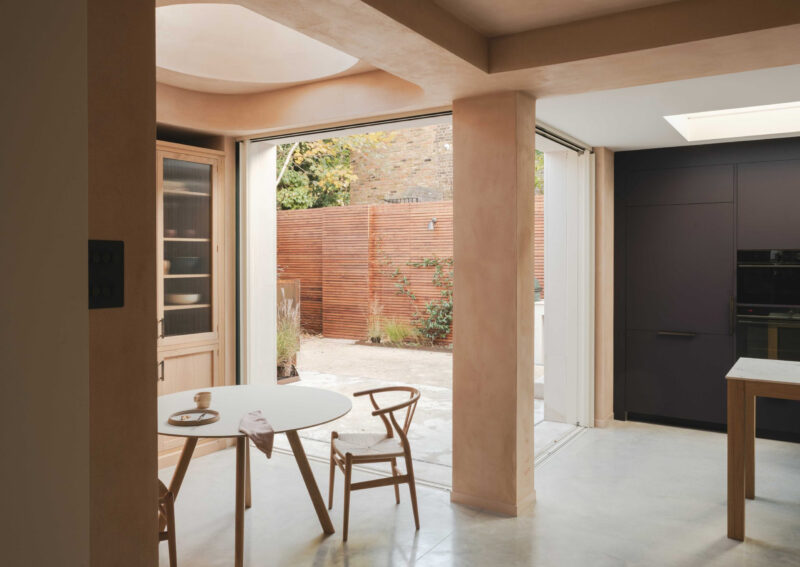Modern open-plan dining area with clay plaster walls, a circular skylight, and a views of a timber-fenced garden.