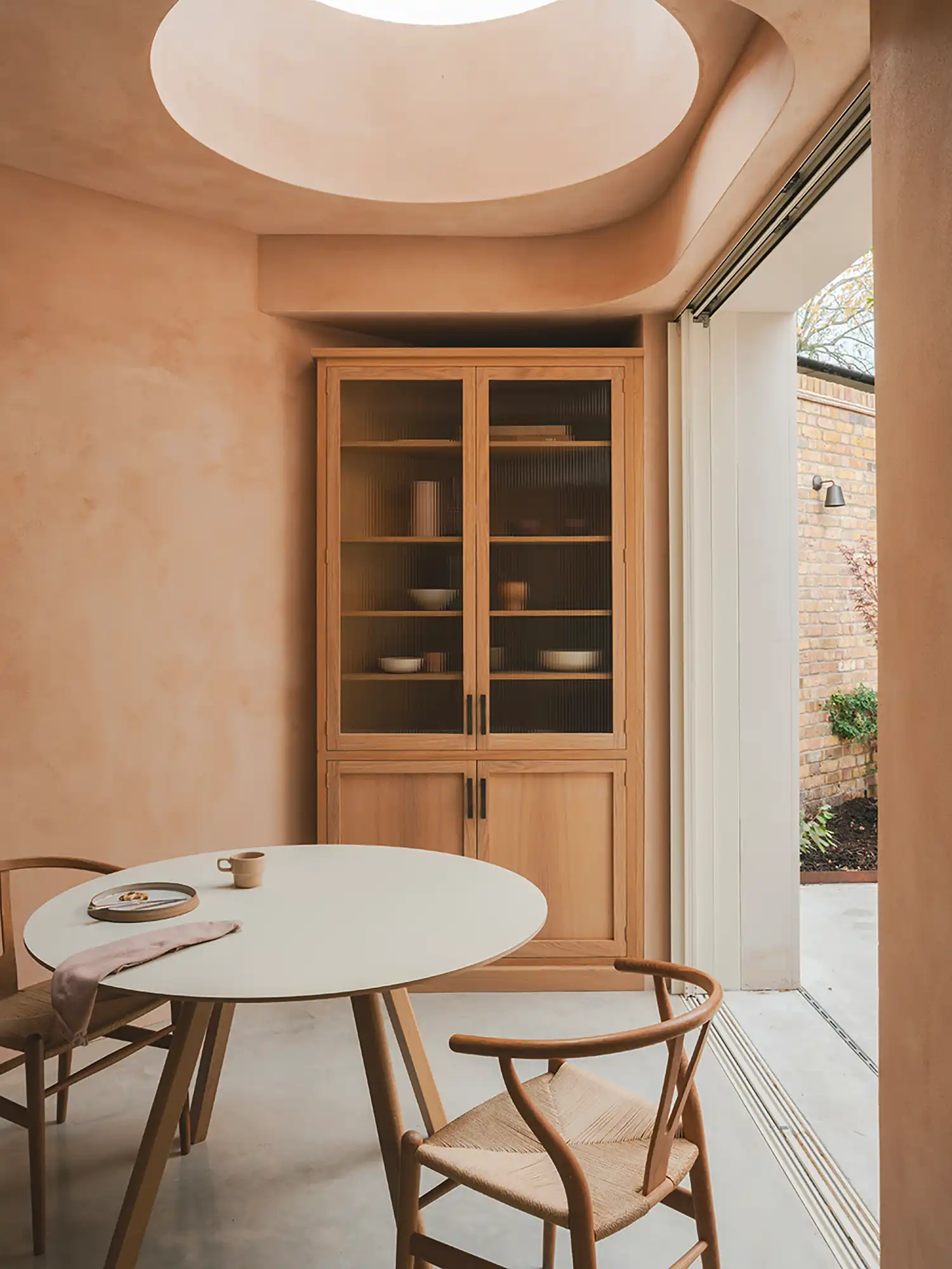 Dining space with a circular ceiling opening, clay plaster walls, and a tall bespoke timber cabinet with fluted glass.