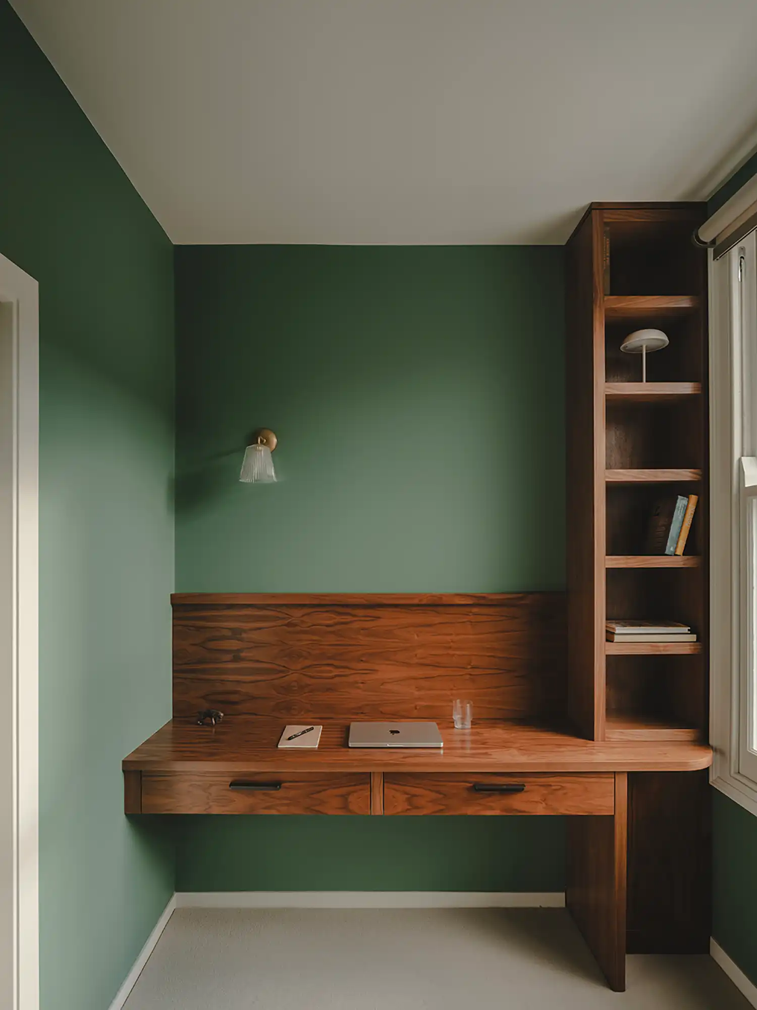 Home office nook with sage green walls, a floating timber desk, and a vertical bookshelf next to a window.