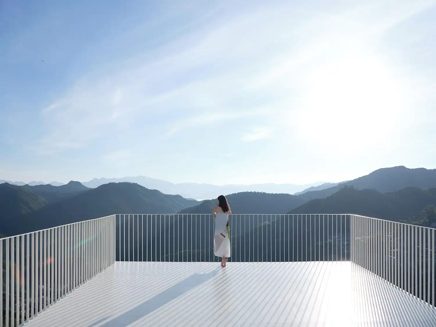 A person standing at the edge of the pavilion’s cantilevered white deck facing a bright, hazy horizon.