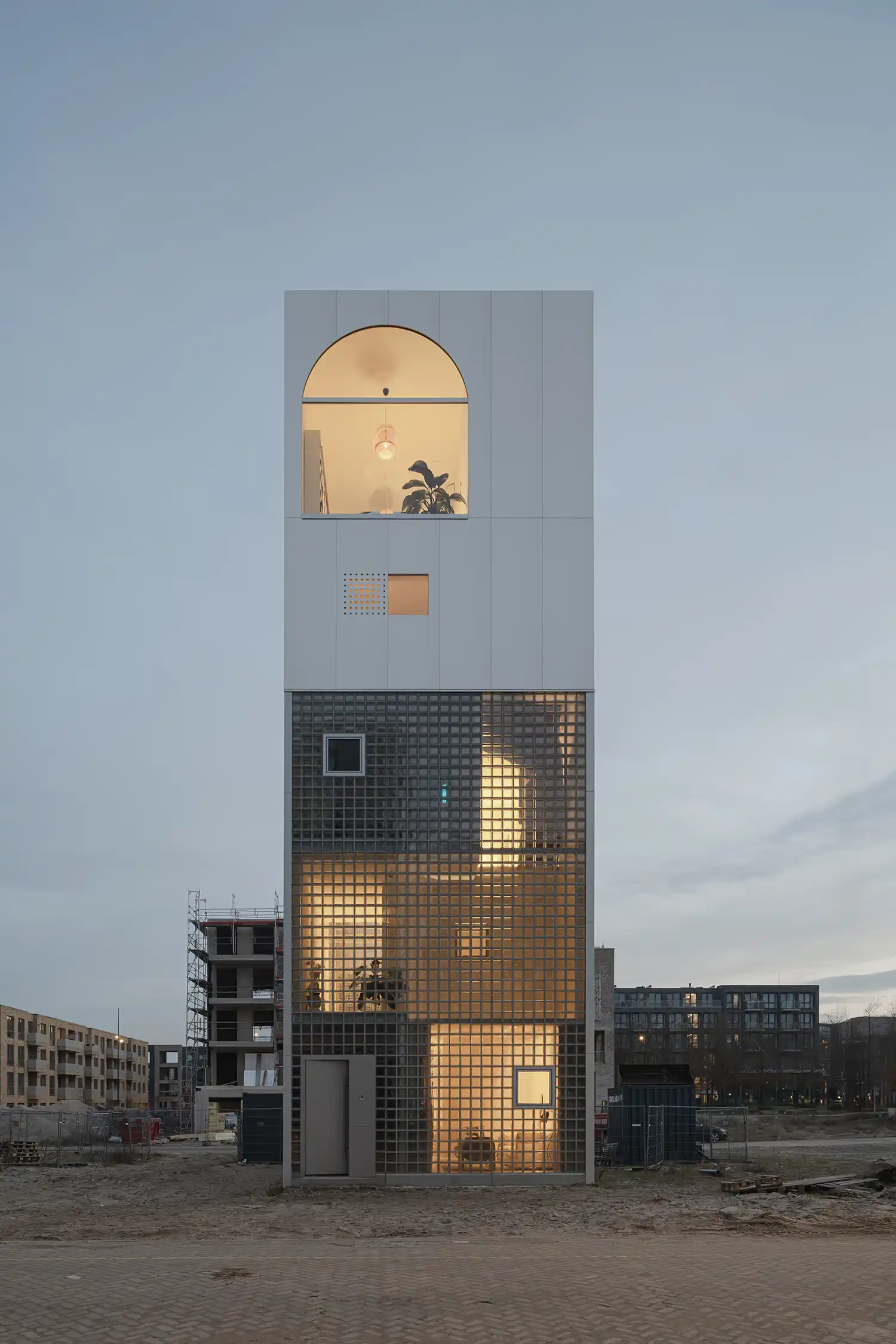 Tall, narrow white and glass building at dusk on a construction site.