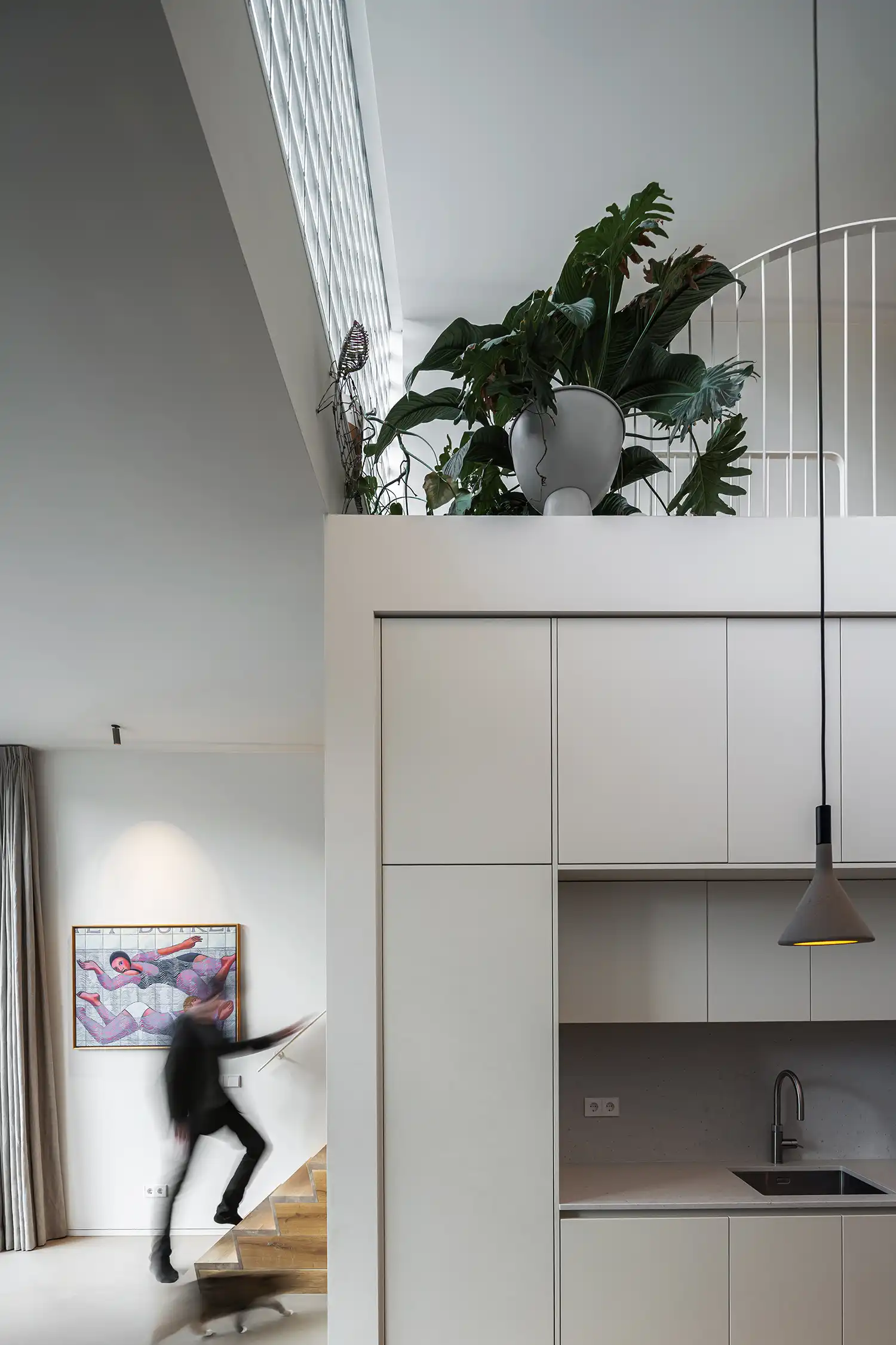 Interior view of a white kitchen with a wooden staircase and a large plant on a mezzanine.