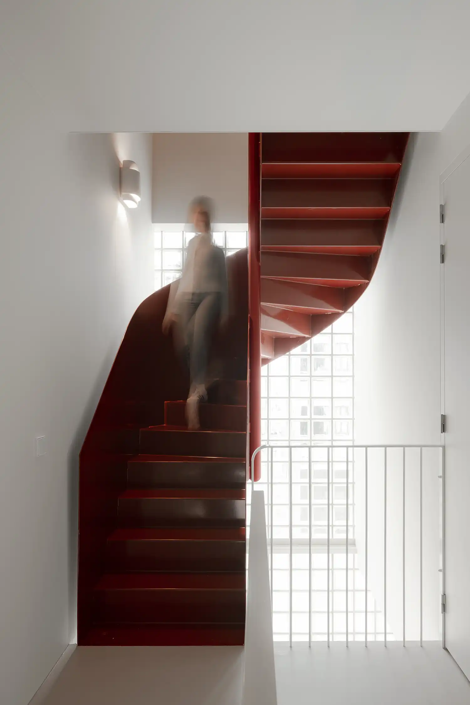 A sculptural red spiral staircase illuminated by natural light from a glass block window.