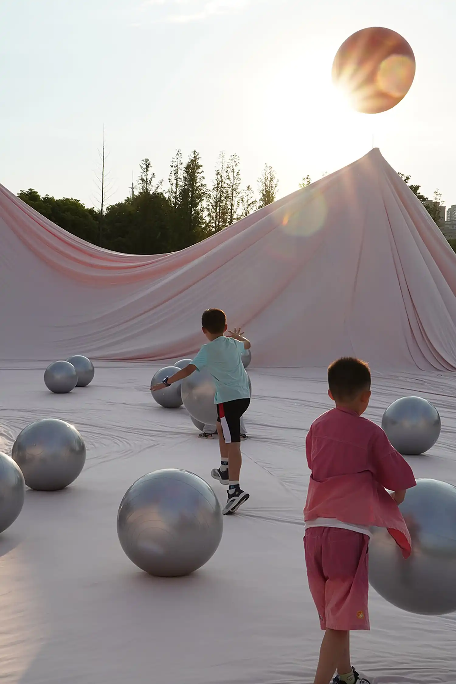 Children playing with silver spheres on a pink fabric surface during sunset.