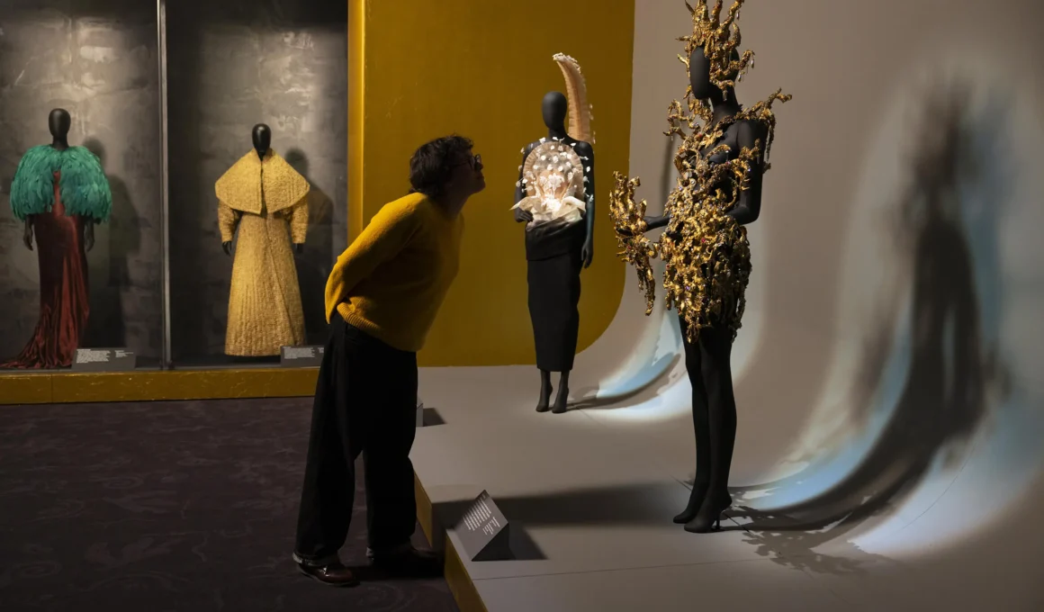 A visitor in a yellow sweater leans in to inspect a gold, sun-ray inspired Schiaparelli sculptural garment on a black mannequin.