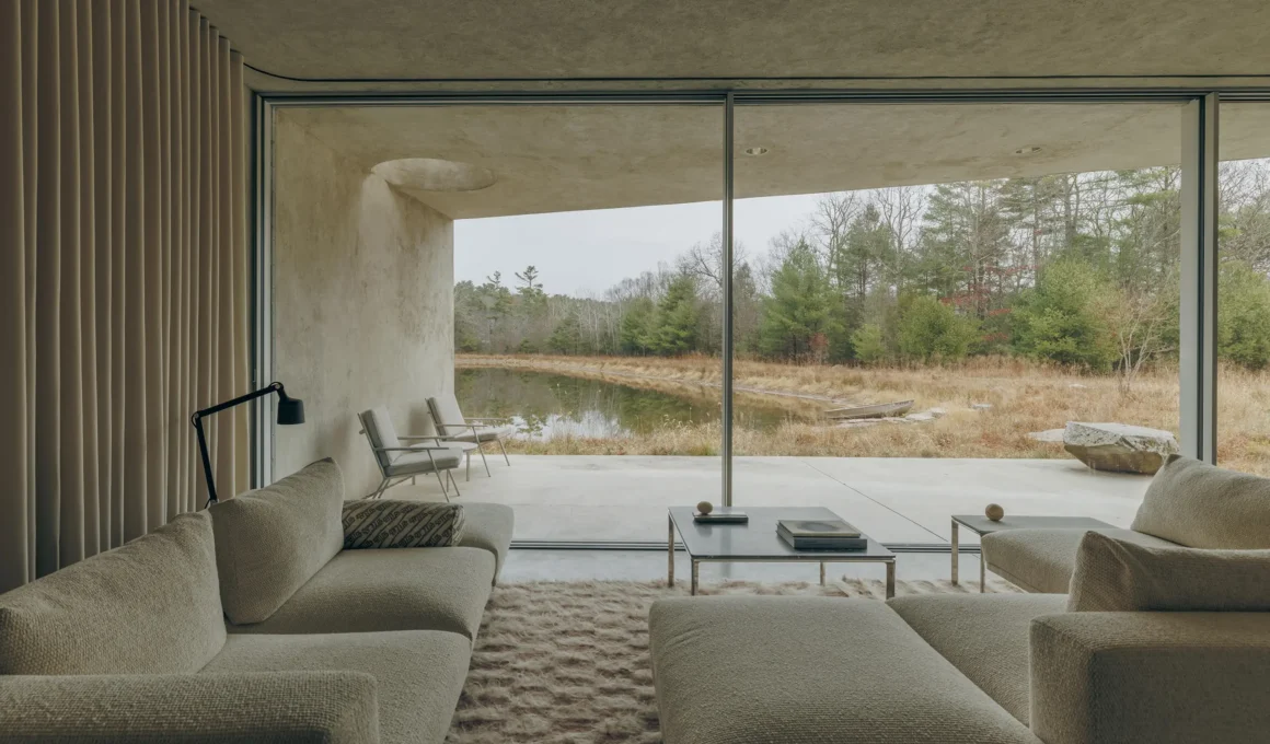 Minimalist living room with beige sofas and a floor-to-ceiling glass wall overlooking a pond in Upstate New York.