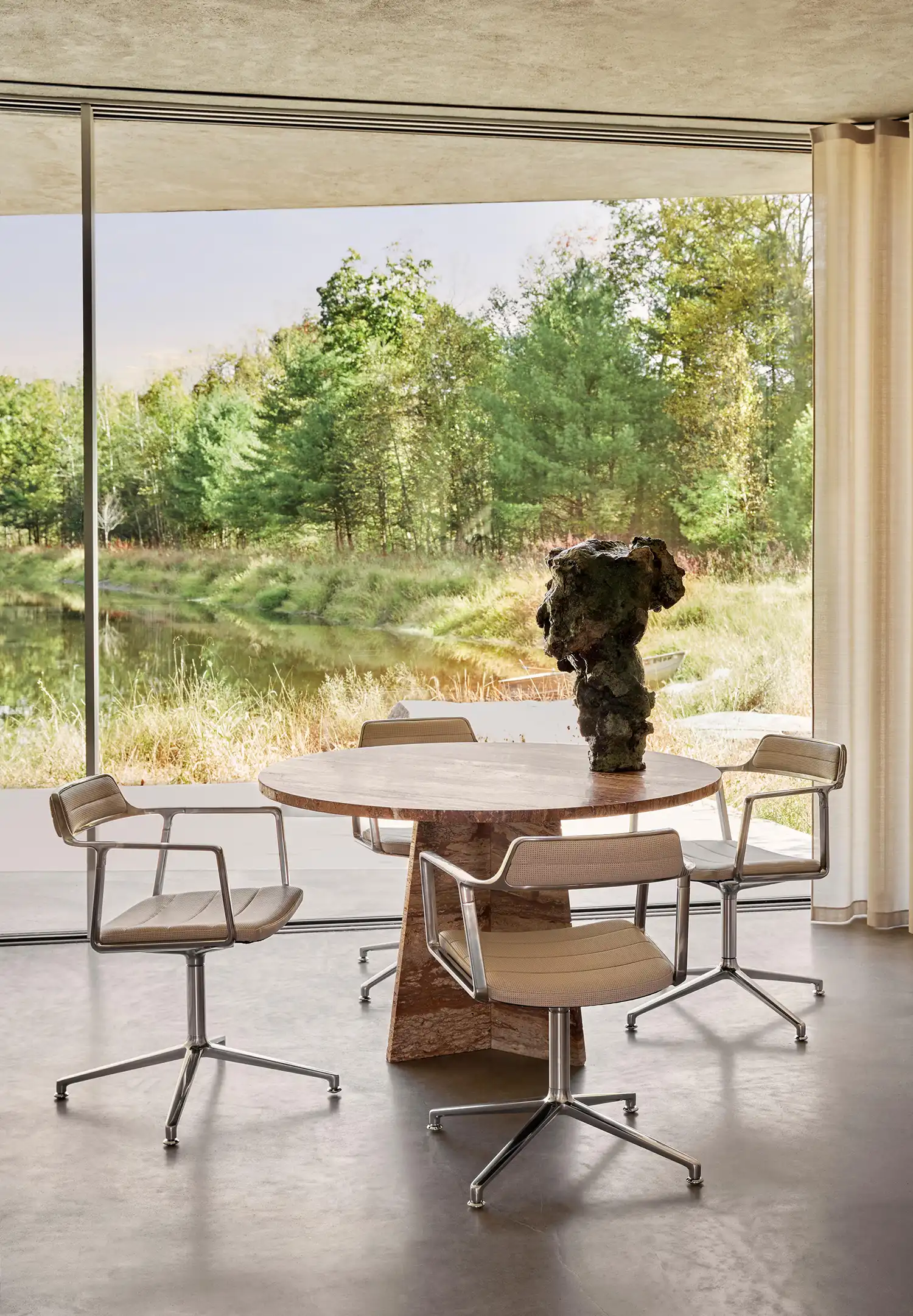 Round travertine dining table and metal Vipp swivel chairs in front of a panoramic window view.