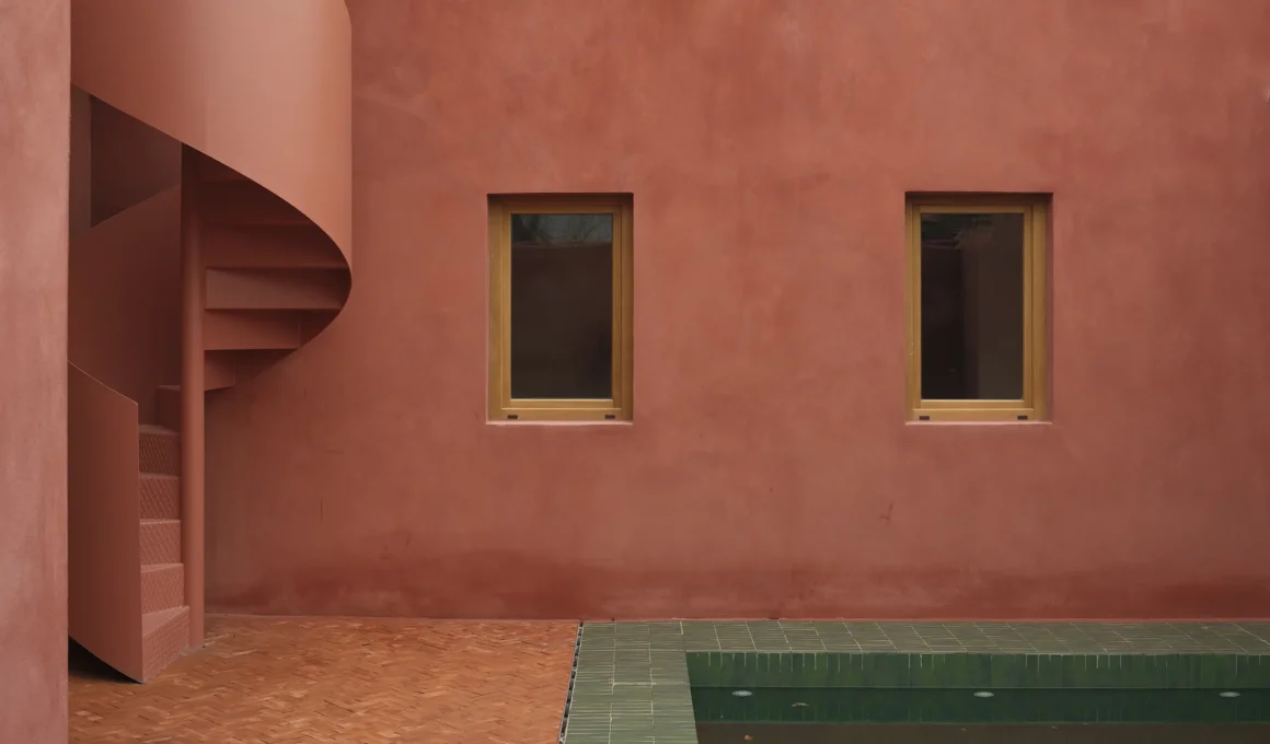 Exterior detail of La Casa de la Charca Verde featuring a terracotta-red wall, a spiral staircase, and the edge of a green tiled pool.
