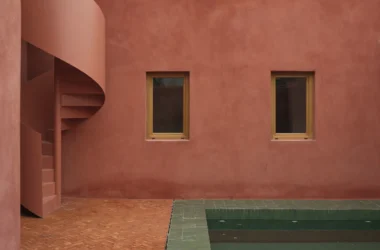 Exterior detail of La Casa de la Charca Verde featuring a terracotta-red wall, a spiral staircase, and the edge of a green tiled pool.