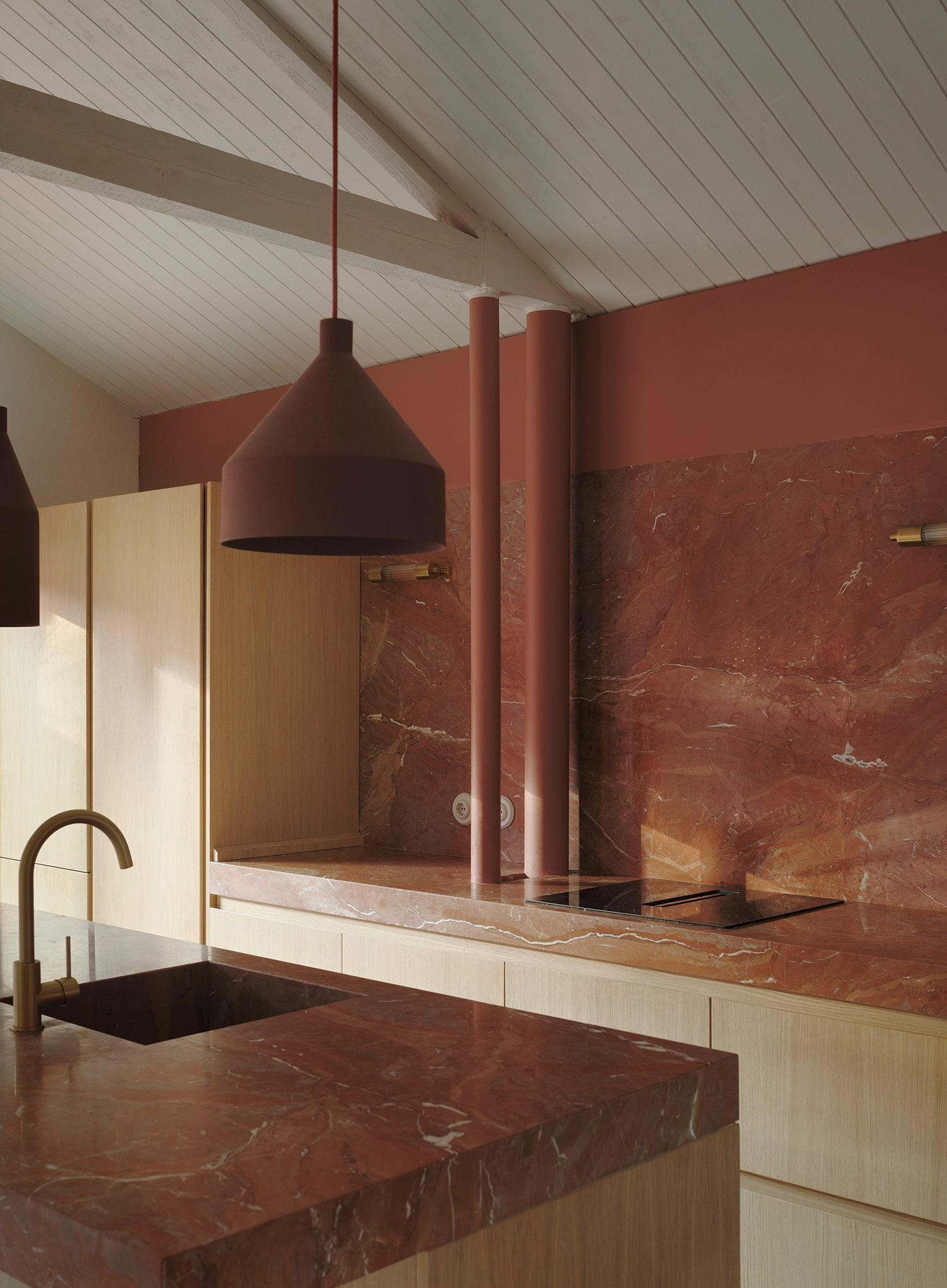 Modern kitchen interior with red marble countertops, light wood cabinetry, and minimalist pendant lighting under a white sloped ceiling.