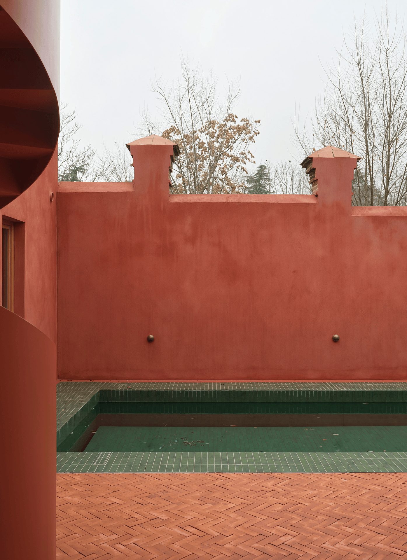 Wide shot of the sunken green pool area surrounded by high red walls and terracotta herringbone paving.