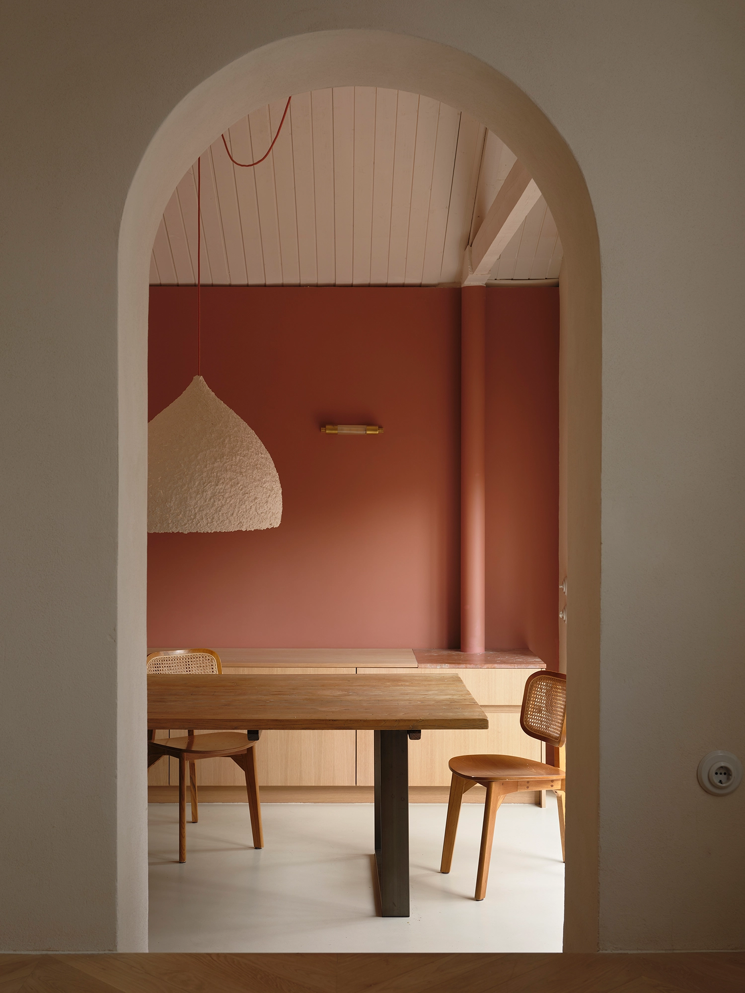View through an arched doorway into a minimalist dining room with a wooden table, woven chairs, and a textured white pendant lamp.