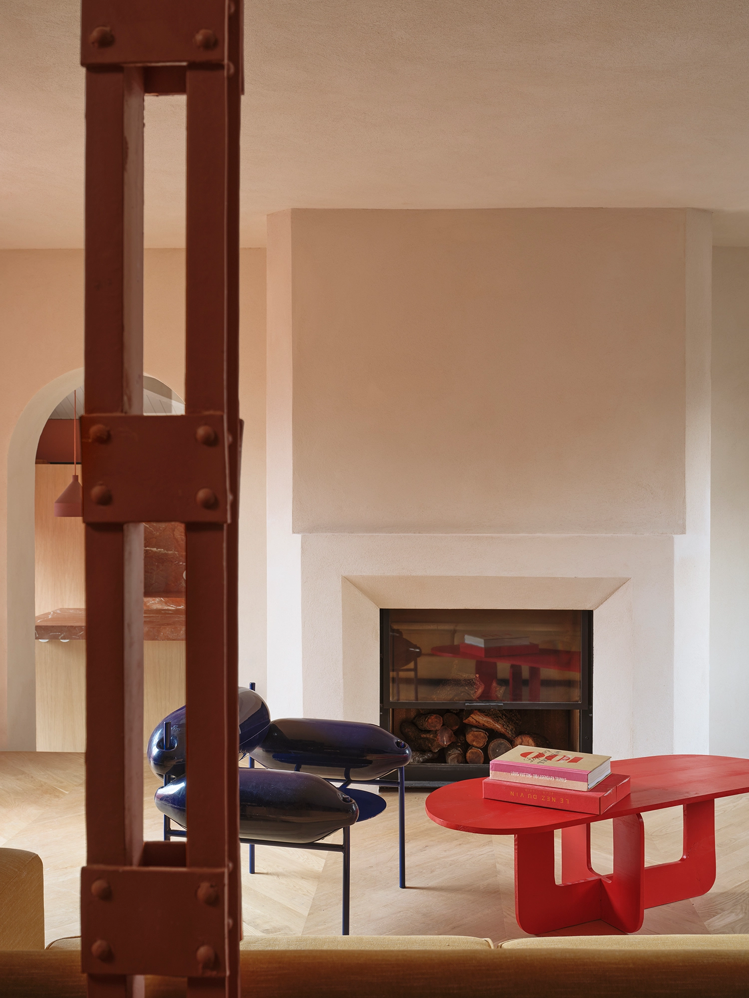 Living room interior with a white fireplace, blue sculptural armchairs, and a red coffee table behind a red structural beam.