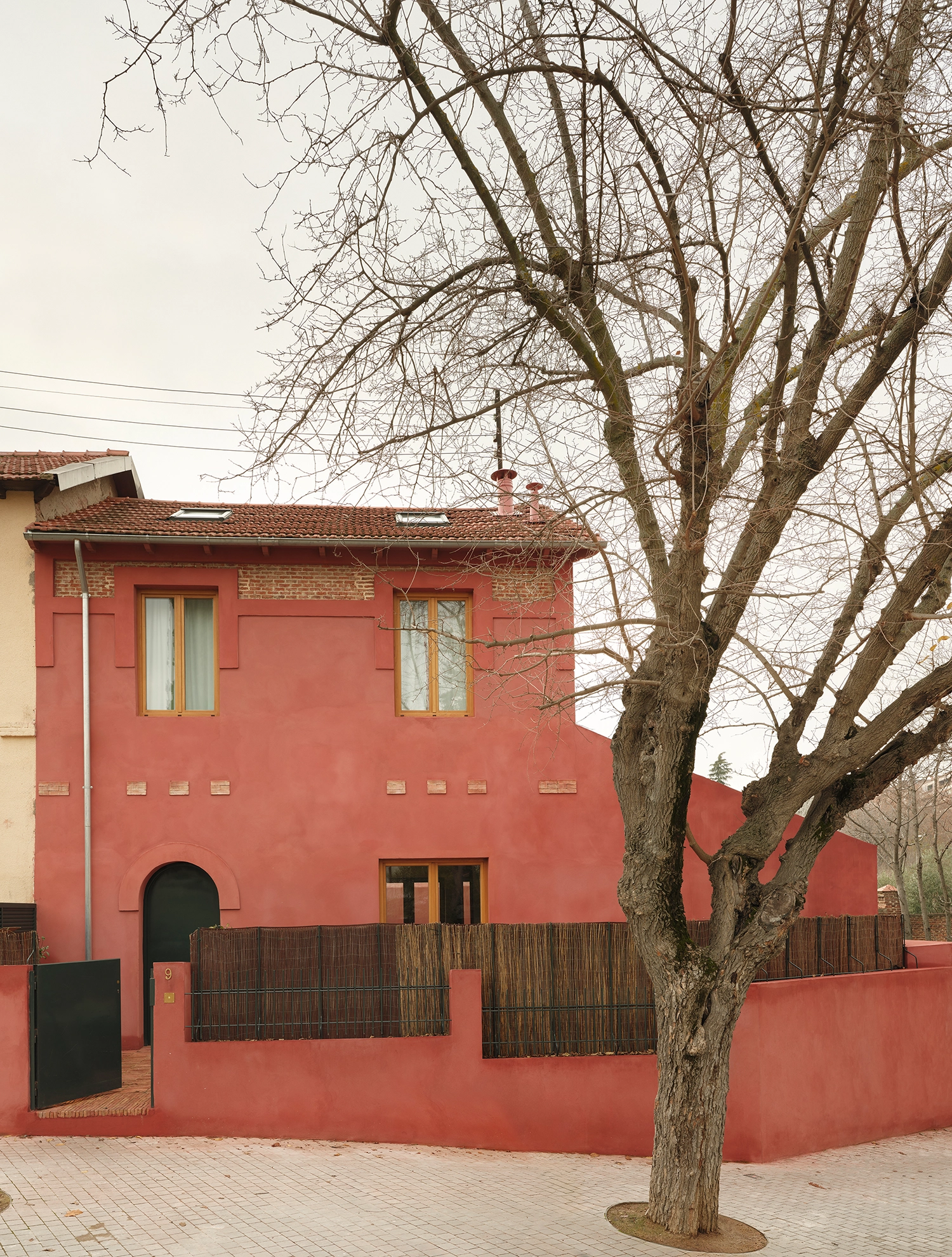 Front exterior of the red-washed Madrid house with an arched green door and a large deciduous tree in the foreground.
