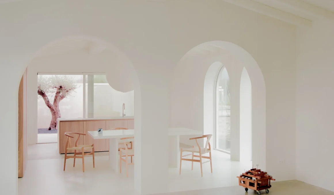 Interior of Casa GA with two white dining tables, several Hans Wegner-style wishbone chairs, and a small olive tree in a courtyard, viewed through white arched doorways.