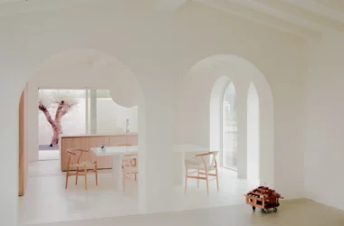 Interior of Casa GA with two white dining tables, several Hans Wegner-style wishbone chairs, and a small olive tree in a courtyard, viewed through white arched doorways.