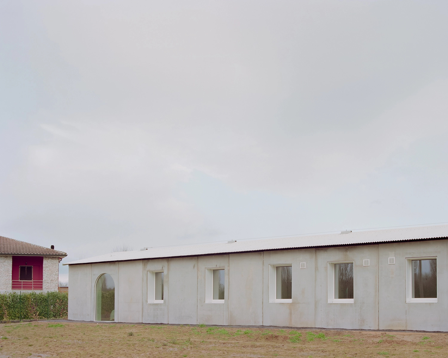 Exterior of a single-story rectangular concrete house with a corrugated white metal roof, multiple square windows, and a tall arched glass door, all set on rough ground.