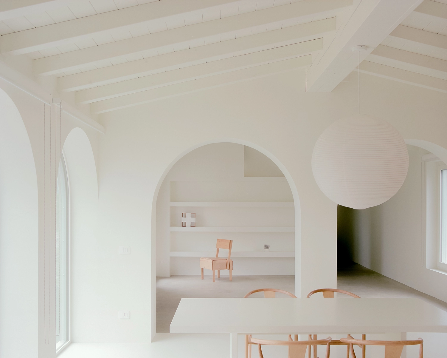 A simple interior room with a white plastered, sloped ceiling, exposed white beams, a round spherical paper lamp, a single wooden chair, and white wall shelving.