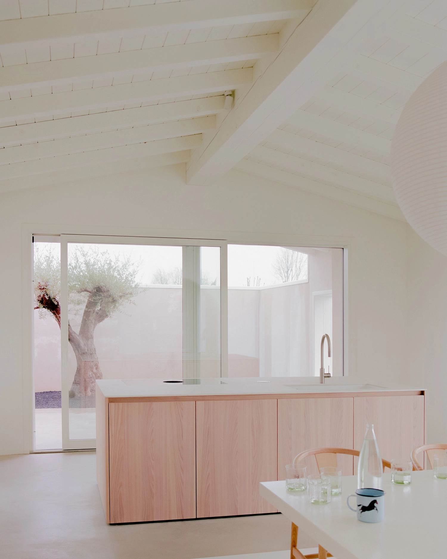 Close-up view in Casa GA of a long, light wood (oak or similar) kitchen island with integrated sink and stovetop, facing a large sliding glass window with an olive tree on a patio.