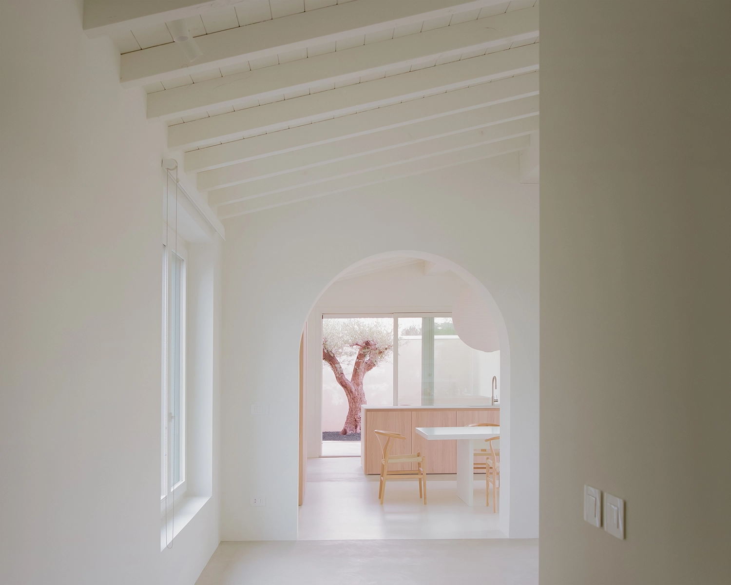 Perspective view through multiple deep white arched doorways, showing a dining area, a light wood kitchen island, and an olive tree patio at the far end, with a framed view on the left.