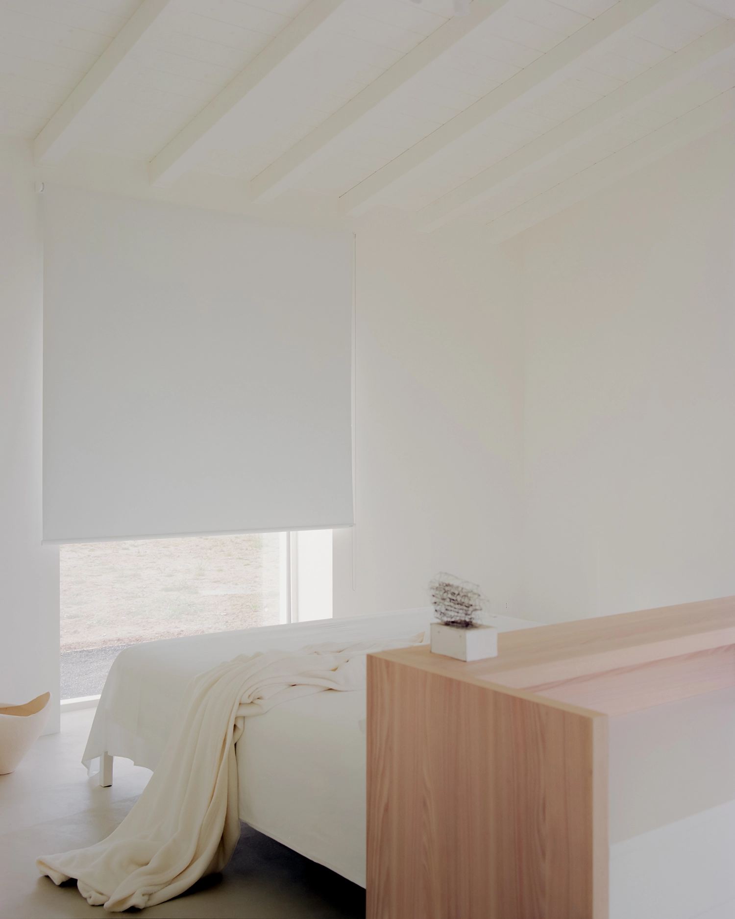 A minimal bedroom with a white bed, neutral blanket, a full-height white window with a roller shade, and a low, simple light-wood partition wall.