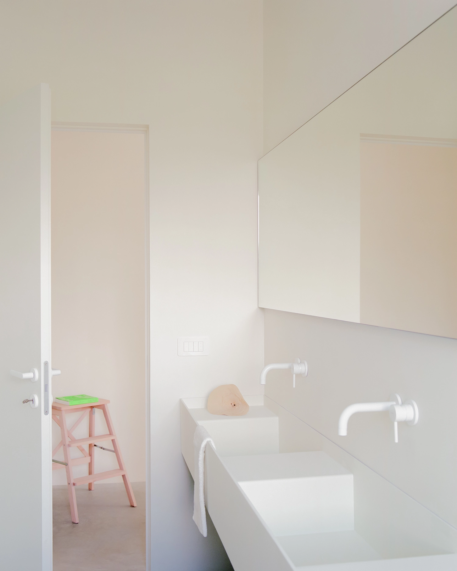 A bright white bathroom corner with a minimal, custom-cast double trough sink, two white wall-mounted faucets, and a large mirror. Through an open door, a pink step stool and a book are visible.