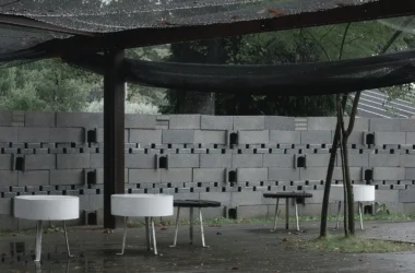 A ground-level view inside the circular Rotação pavilion by Atelier Tiago Antero, focusing on the curved, textured concrete block wall and the minimalist white and black stools placed on the wet pavement.