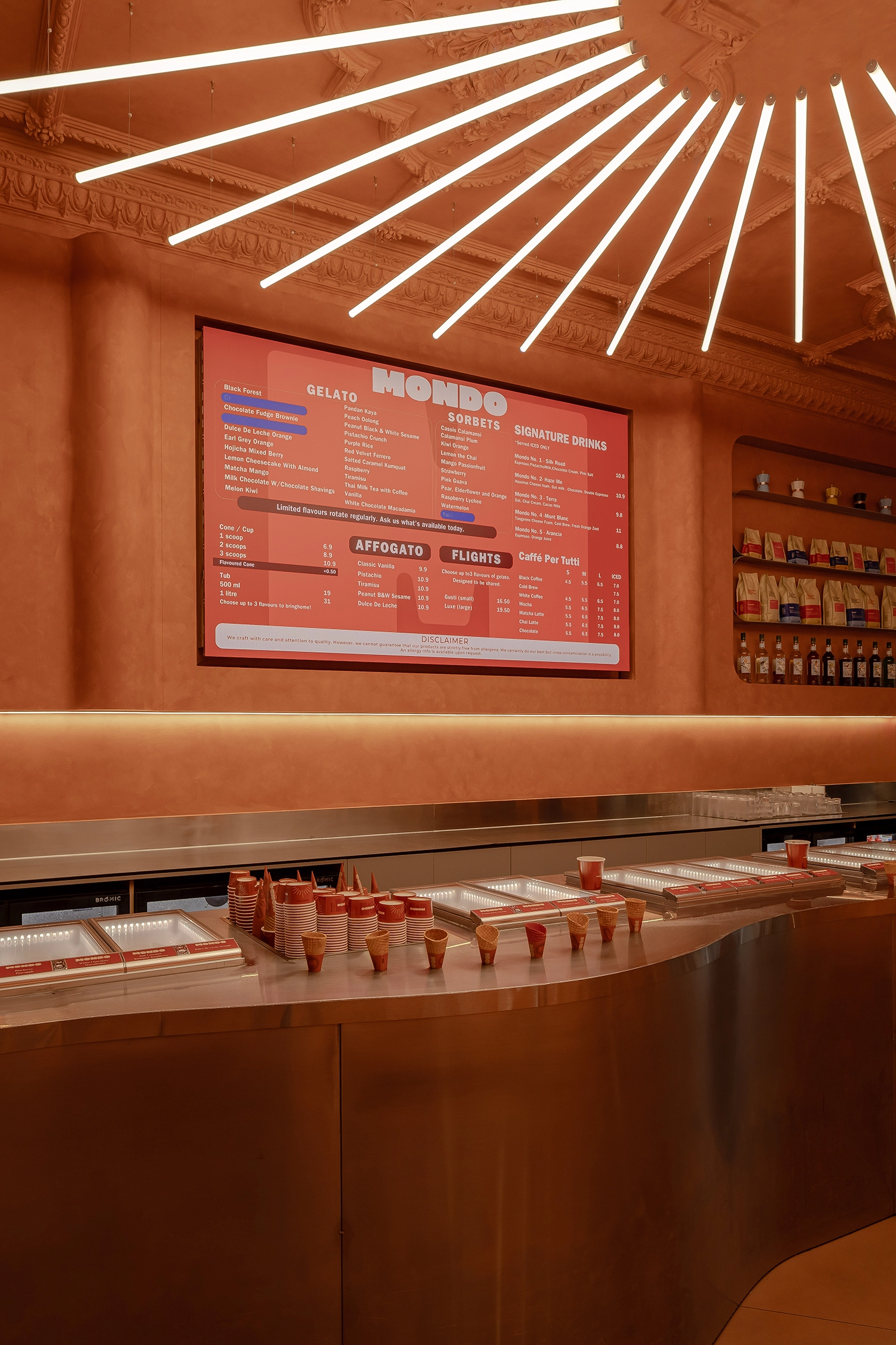 Close-up of a brushed metal service counter with embedded gelato displays under a glowing digital menu board.