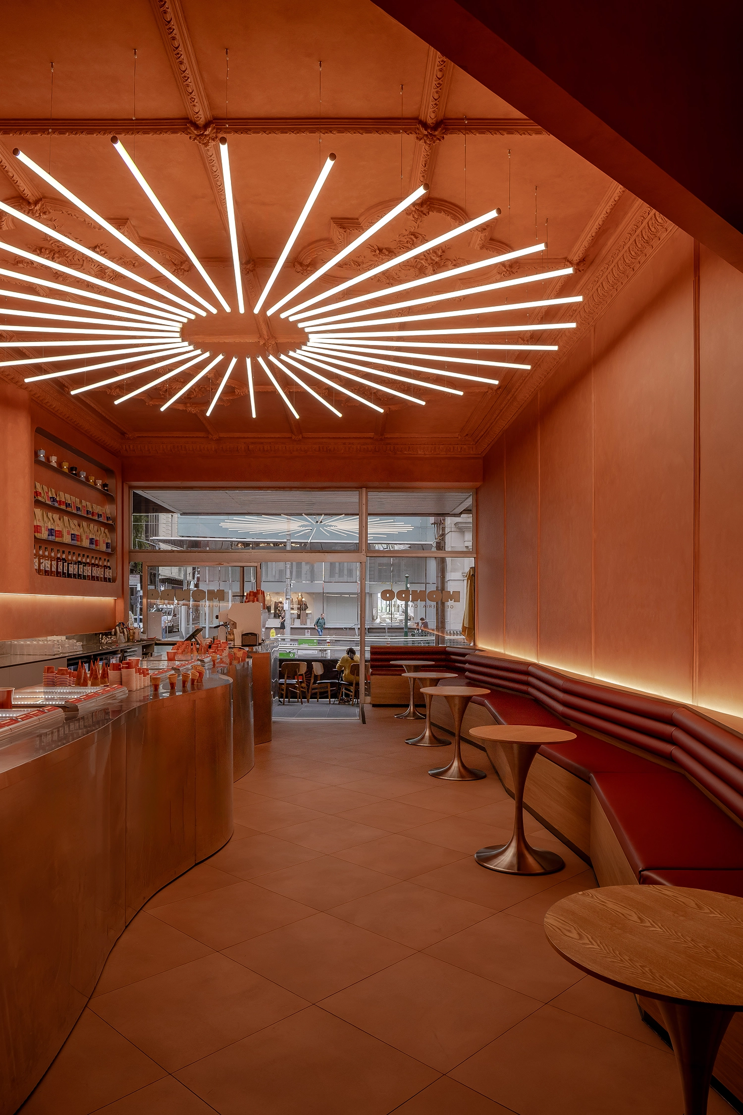 Perspective view from the back of the gelateria looking toward the street, showing the long burgundy banquette and sun-burst light.