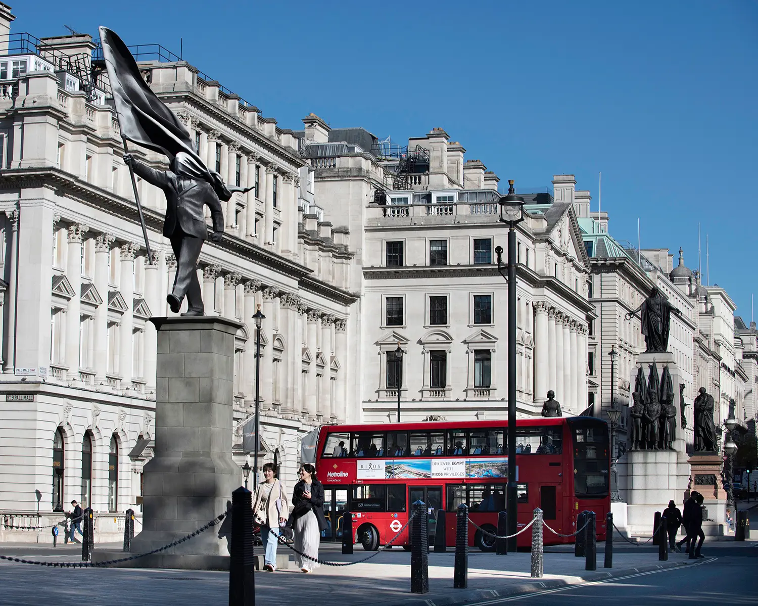 Wide shot of Banksy's sculpture in Waterloo Place, London, with a red double-decker bus and St. James architecture.