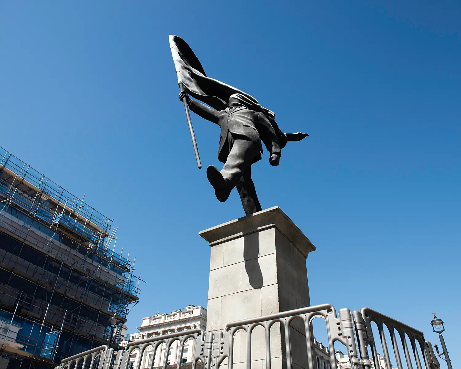 Low-angle view of the Anonymous Flag-Bearer statue by Banksy on a pedestal in London, surrounded by security barriers.