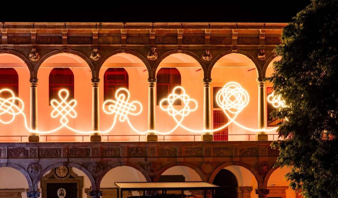 Illuminated LED knots by BIG and Artemide glowing against the Renaissance arches of the University of Milan at night.