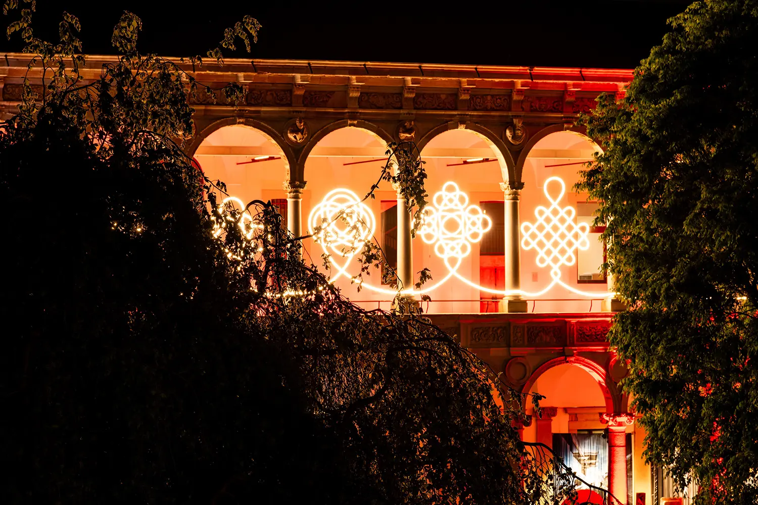 Warm LED knots glowing behind silhouettes of trees at the University of Milan during a FuoriSalone night event.