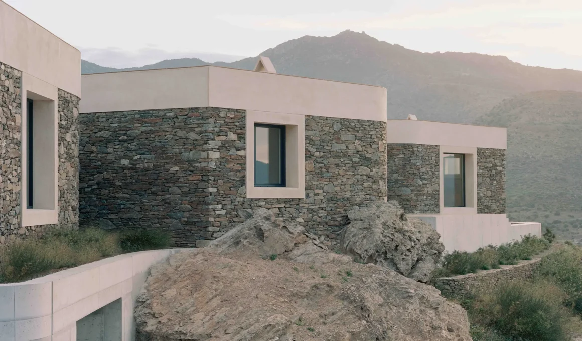 Exterior view of multiple cubic volumes with textured stone walls and smooth concrete caps cascaded against natural rock formations and a mountain backdrop under soft light.