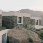 Exterior view of multiple cubic volumes with textured stone walls and smooth concrete caps cascaded against natural rock formations and a mountain backdrop under soft light.