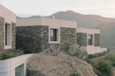 Exterior view of multiple cubic volumes with textured stone walls and smooth concrete caps cascaded against natural rock formations and a mountain backdrop under soft light.