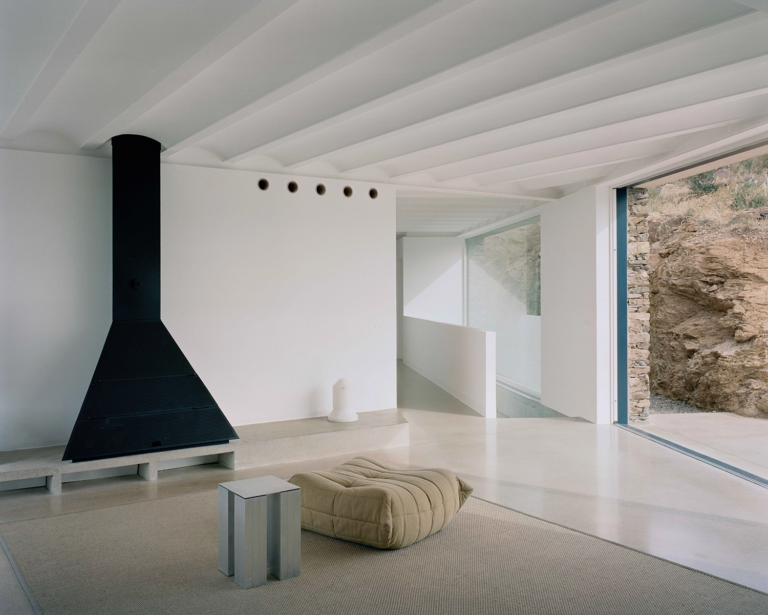 Minimalist interior living room with a geometric black metal fireplace, white beamed ceiling, textured beige ottoman, and large glass door opening to a rocky outcrop.