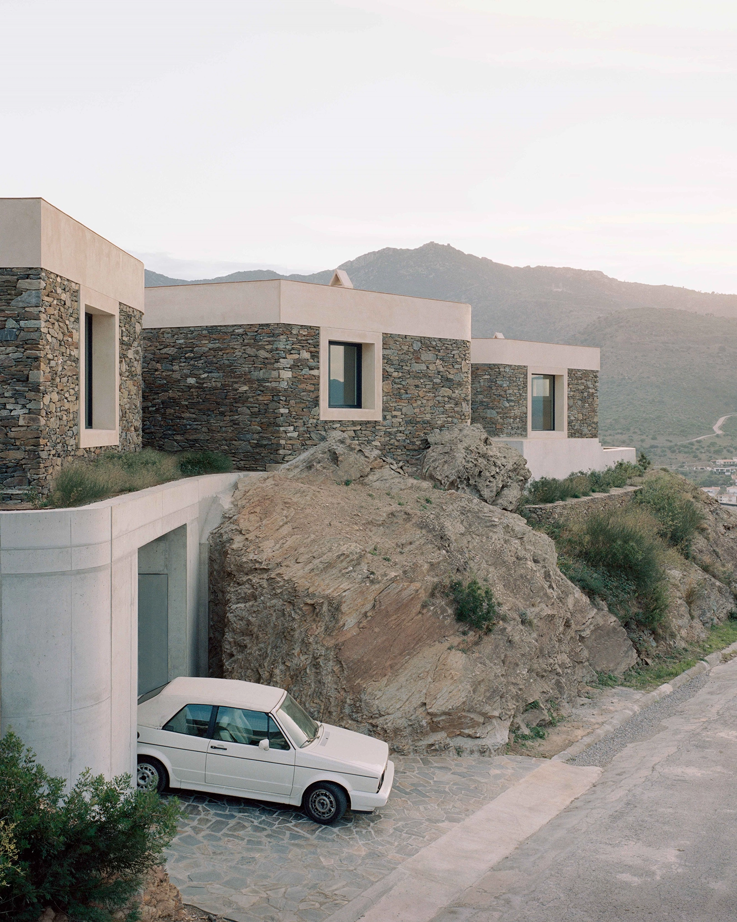 Exterior view of stacked stone and concrete volumes on a hillside, with a vintage white convertible car parked in a lower concrete garage next to large natural boulders and mountains in the background.