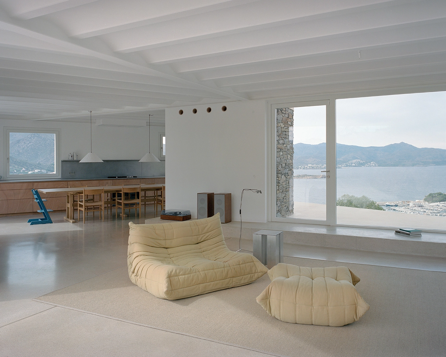 Minimalist open-plan interior living and dining area with white vaulted ceiling and expansive sea view through large sliding glass doors, featuring wooden furniture and a beige ottoman.