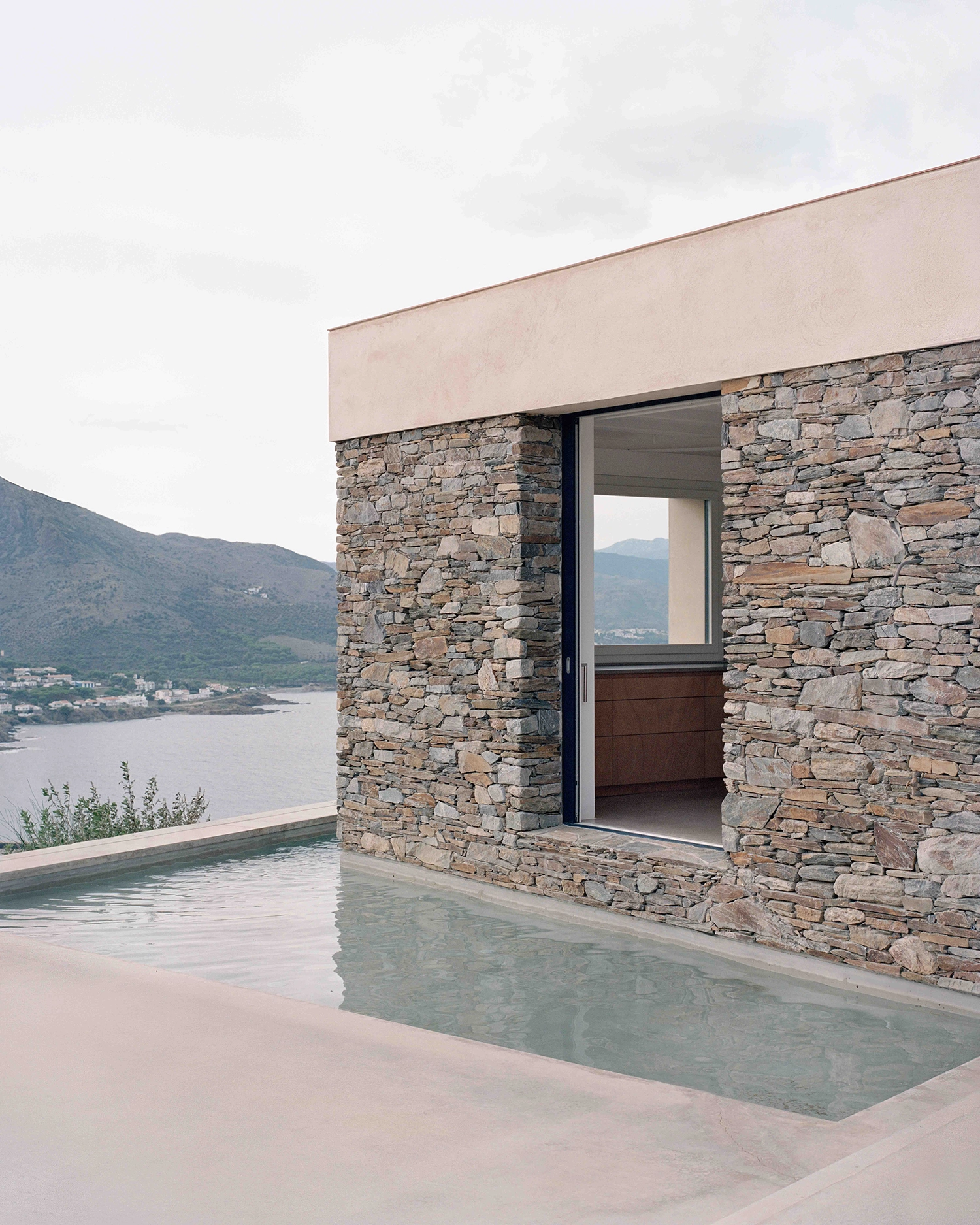 Exterior corner of Casa Balma Murada featuring rough stone walls, a smooth concrete deck, and a narrow swimming pool with a panoramic view of the sea and distant mountains under an overcast sky.