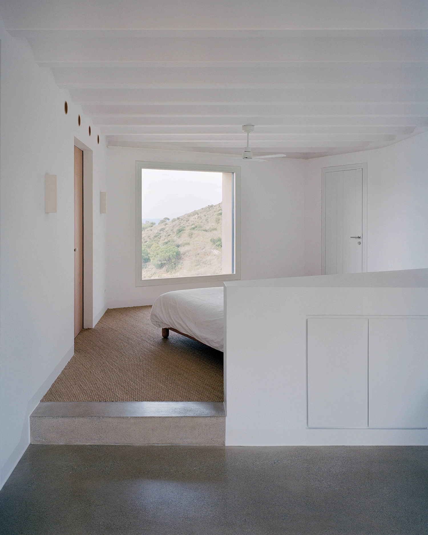 Minimalist bedroom interior with white walls, a vaulted ceiling, a low bed, and a large square window framing a rolling arid hillside view.