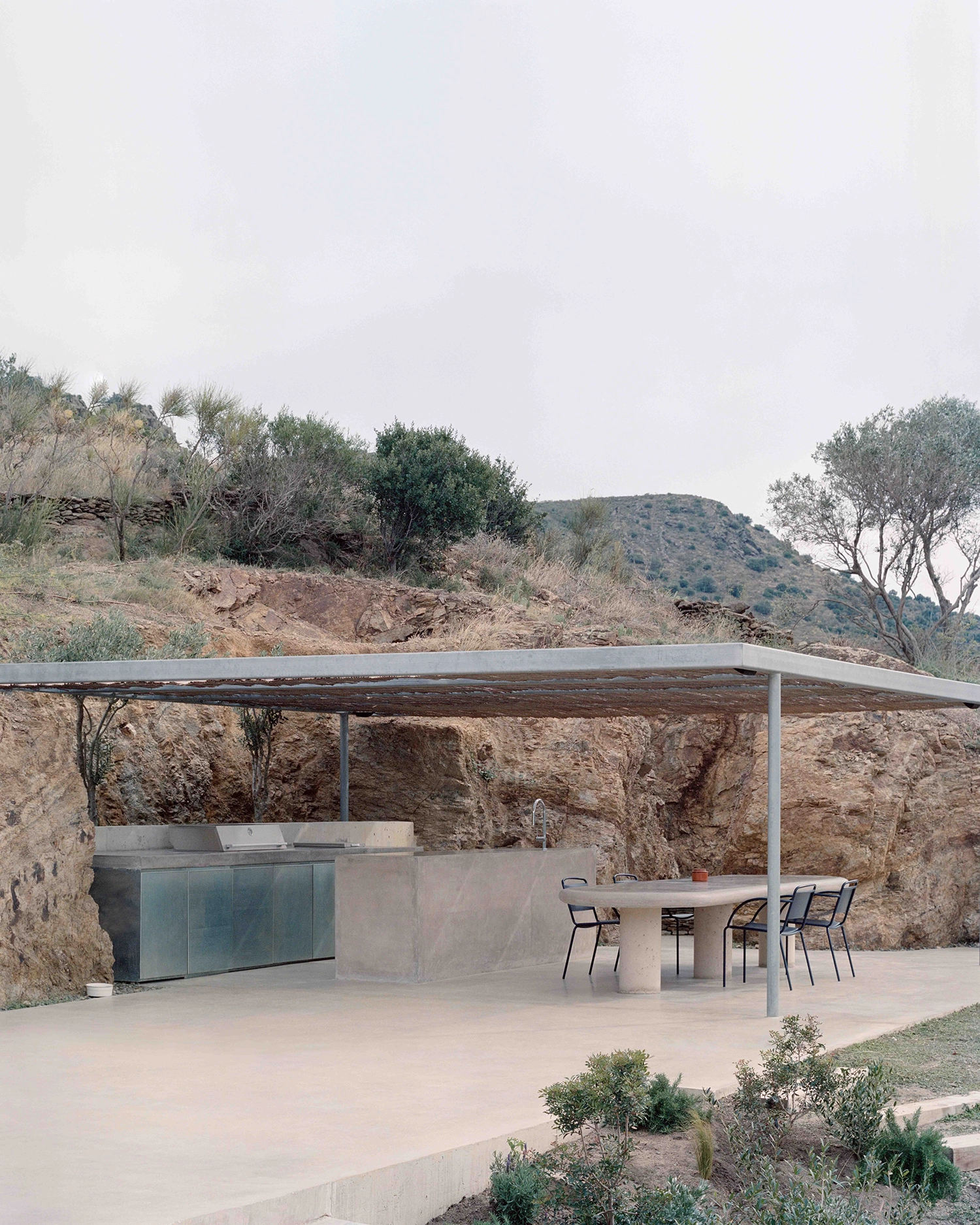Outdoor concrete kitchen and dining area under a minimalist steel and trellis shelter, set against a raw rocky bank on a hillside.