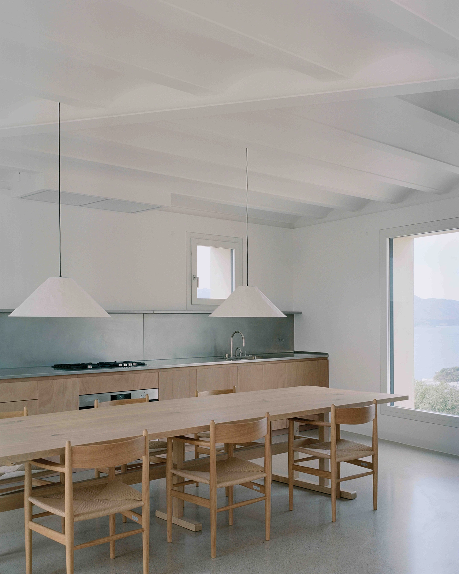 Minimalist interior kitchen and dining area with light wood cabinets, stainless steel countertop, white vaulted ceiling, two geometric paper pendant lamps, and a sea view window.