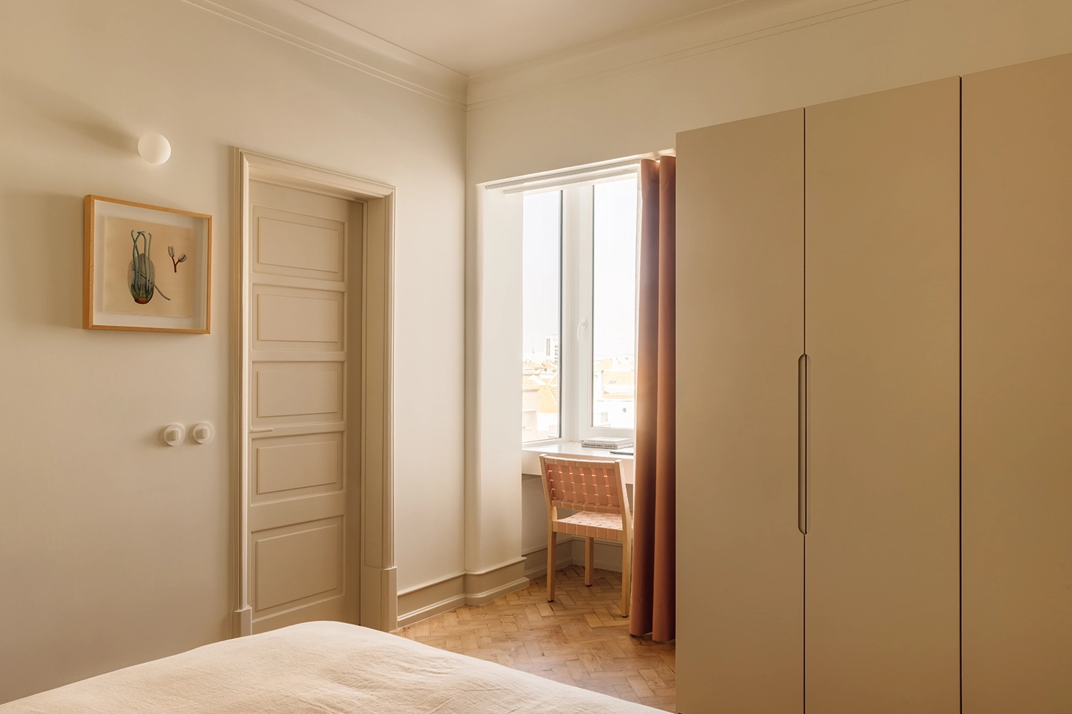 Minimalist bedroom in Lisbon with herringbone parquet flooring, cream-colored cabinetry, a framed botanical print, and a pink curtain next to a window view.