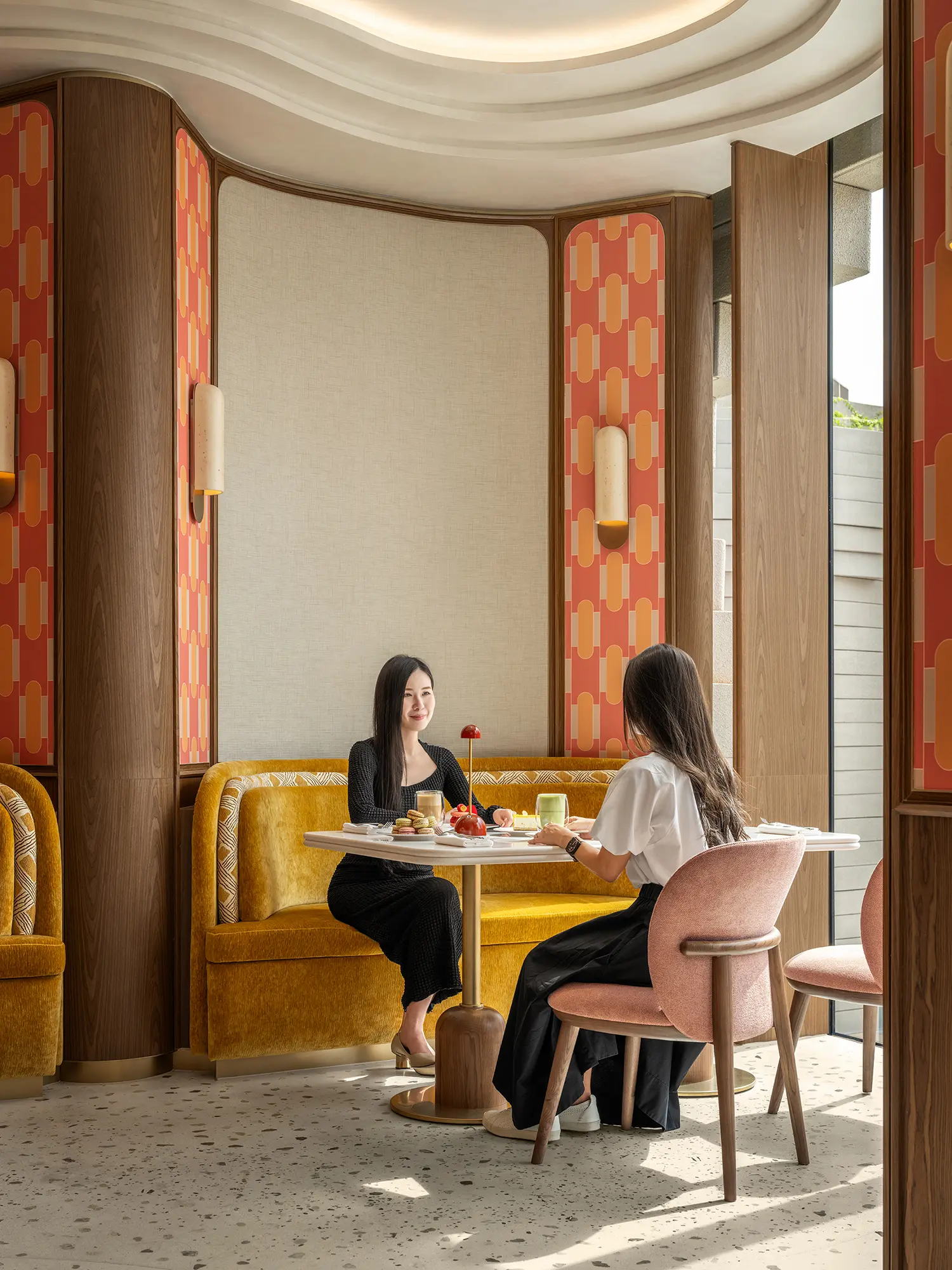 Two women seated at a marble table in a booth with yellow velvet seating and patterned wallpaper.