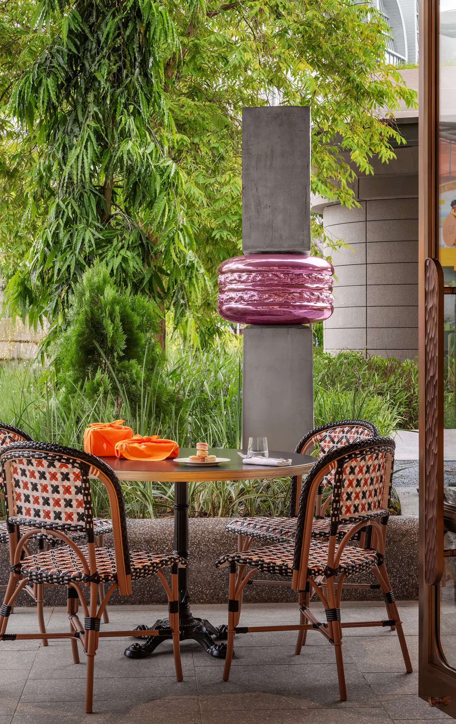 Outdoor seating area with Parisian-style chairs and a large pink macaron sculpture among greenery.