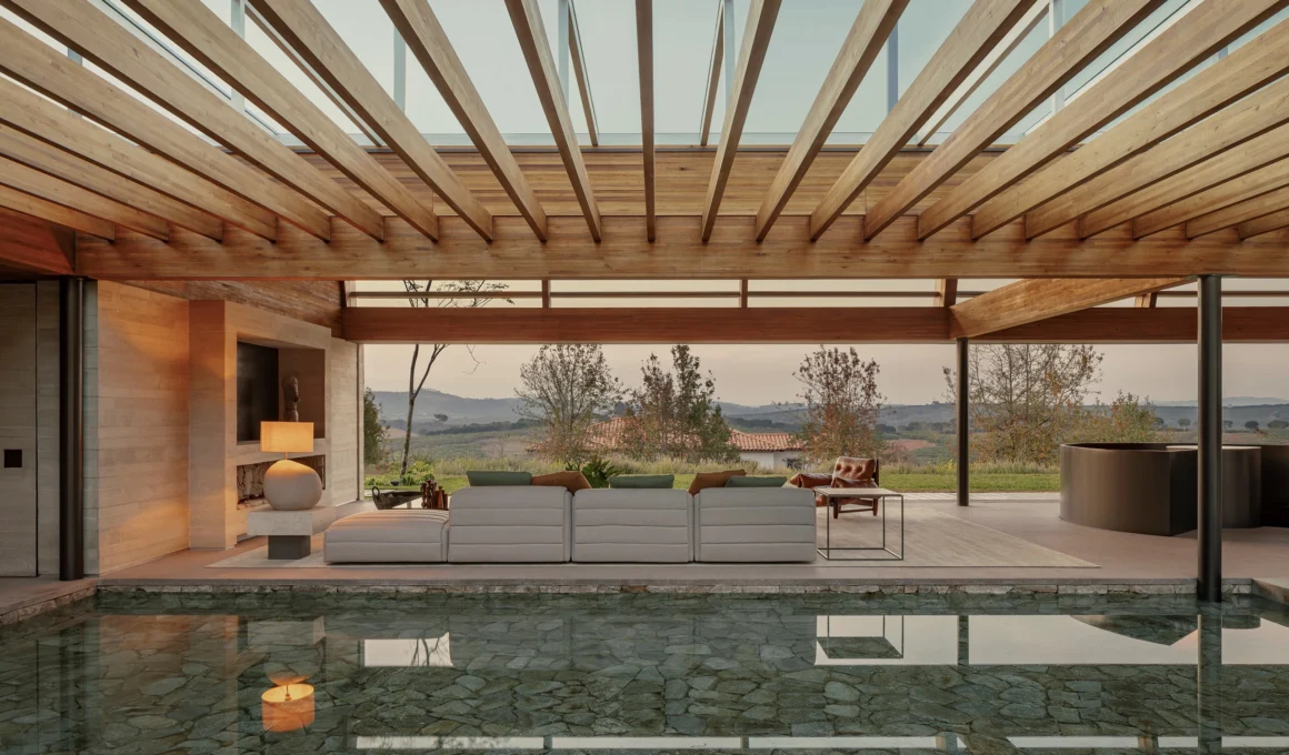 Wide-angle architectural photograph of a seamless indoor-outdoor transition between a luxury lounge and a flush swimming pool, showing a deep perspective through a louvered wooden ceiling structure.