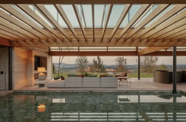 Wide-angle architectural photograph of a seamless indoor-outdoor transition between a luxury lounge and a flush swimming pool, showing a deep perspective through a louvered wooden ceiling structure.